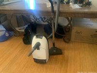 Image of Miele White Star vacuum cleaner with hose and brush head positioned on wooden floor near table.