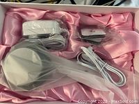 Handheld treatment device, power adapter, cable, and small attachment in a pink satin-lined box, all sealed in plastic.