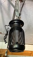 Bug zapper hanging from a metal hook showing the protective black plastic grill and a power cord with standard plug.