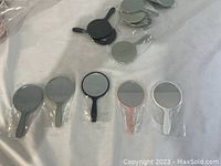 Photo showing five mini handheld mirrors of different colors laid out with their handles visible, packaged in plastic wrapping.