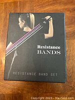 Photo of the front of the resistance band set box showing a woman using the bands for exercise. The box is black with an image of the bands in use, including different colored bands illustrating multiple resistance levels.