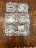 Six rolls of double sided adhesive tape, sealed in clear plastic, arranged in two columns on wooden floor.