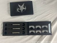 Image shows opened black foam-padded box with three steel-tip darts secured on left side and six arrow-shaped black flight fins with white and silver chevron design on right side, black box top with white star emblem.