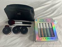 Makeup bag, three e.l.f. powder jars, two makeup brushes, and a boxed 5-piece glitter lip transformers set arranged on white fabric.