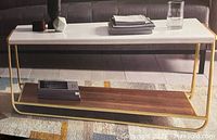 Photo of the assembled Sayer Coffee Table showing white top, gold frame, and lower wood shelf in a living room setup