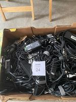 Box filled with a variety of black power adapters and chargers tangled together, showing different sizes and types of cords.