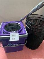 Photo showing purple spin mop bucket with mop handle and black mesh wastebasket.