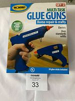 Front view of sealed package showing two blue Ideaworks glue guns and text highlighting multi-task usage, home repair, crafts, and included 20 glue sticks.