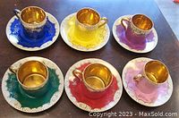 Six colored demi-tasse cups placed on matching saucers with gold lustre interiors and detailed gold accents.