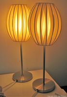 Pair of mid-century modern style table lamps lit, showing oval woven fabric shades with vertical openings and brushed metal bases.