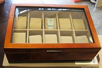 Closed wooden watch display case with glass top showing one watch inside and nine empty suede cushions.