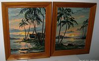 Pair of two oil paint by numbers paintings in oak frames leaning against a white wall, showing tropical beach scenes at sunset with palm trees and huts.