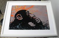 Front angled view of framed Banksy style pensive monkey art print outdoors on concrete surface showing orange splatters and contemplative expression of monkey.