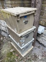 Stack of four large taupe plastic totes with lids, showing wear, paint marks, and dirt, placed outdoors against stone and wood surfaces.