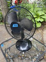 Black 3 speed oscillating table fan placed outdoors on a glass table with greenery in the background