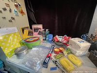 Camping accessories including plastic tablecloths, utensils, bowls, foil, rope, napkins, all arranged on a table.