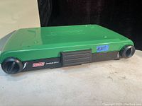 Green Coleman double burner propane stove, lid closed, showing front with black knobs and Coleman branding.