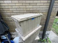 Two beige plastic storage totes stacked outdoors against a brick wall, showing lids with securing clasps and normal age wear.