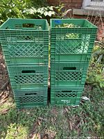 Six green plastic milk crates stacked outside on grass with a brick wall and plants in the background. The crates have standard milk crate design and visible cut-out handles.