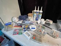 Photo of the complete lot showing various hardware items on a white table including caulking tubes, paint rollers, burlap roll, rope, and assorted fasteners in containers and packages.