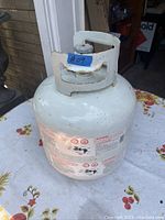 Photo of used white propane BBQ tank with safety and usage labels on the side, placed on floral-patterned table outdoors.