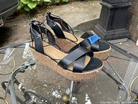 Front and side view of pair of black wedge sandals on glass table outdoors.