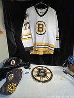 White Boston Bruins hockey jersey with number 73 hanging, black furry trapper hat, baseball cap and large Bruins patch on table.