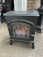 Black electric fireplace heater with simulated burning log effect, standing outdoors on concrete, showing front and top metal surface.