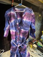 Photo of a folded long house coat in shades of purple, blue, and pink tie-dye. It is wrinkled and hanging on a hanger, placed in a cluttered background with various tools and containers.