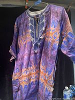 Photo of a traditional Nigerian shirt hanging on a hanger against a black background, showing purple and orange design with white embroidery.