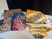 Folded yellow and brown tie-dye Nigerian table cloth and 5 matching napkins arranged beside other colorful folded Nigerian fabric