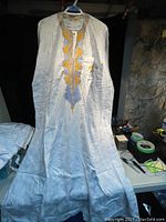 White long-sleeve Nigerian top with yellow and blue embroidery hung on a hanger, displayed front view.