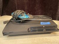 Black Sony PS3 console with a wired translucent blue and black controller on top, console front visible with disc drive and buttons.