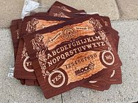 Photo showing a stack of 17 wooden-style mouse pads printed with Ouija board design including alphabet, numbers, YES and NO circles, and decorative elements.