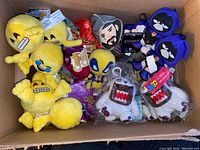 Box containing 33 assorted plush toys including multiple emoji faces with varied expressions, Domo characters, Candy Crush and superhero themed toys such as Deadpool, some with tags and packaging.