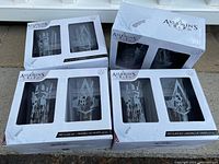 Four boxed sets of two Assassin's Creed pint glasses each, arranged grouped together showing outer packaging with logo and window to see the glasses.