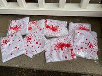 Eight white tea towels with red blood splatter design arranged on the ground, each individually wrapped in plastic.