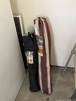 Two carry bags containing the folding camp chairs, one brown with beige stripes, one black, standing against a wall.