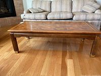 Top and side view of wooden coffee table on hardwood floor in front of couch. Shows wood grain top and brass accents on legs.