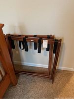 Two wooden luggage racks, one folded against the wall and one upright, both made of medium-tone wood with dark fabric straps for supporting luggage.