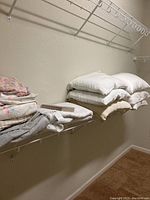 Six standard bed pillows stacked on a shelf with assorted bedding linens underneath.