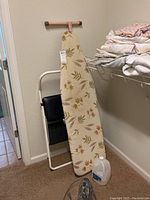 Photo of ironing board leaning against wall with fabric cover showing beige background with leaf pattern, part of the two-step stool visible behind.