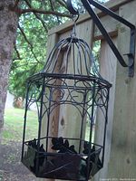 Front angle view of a black metal cage-style lantern with hinged lid, hanging from a bracket on a wooden fence, outdoor setting with trees.