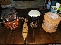 Overview of four drums including the tall carved wooden drum with animal skin top, birch bark drum with suede top and bottom, black and white hand carved drum, and Acapulco handmade instrument.