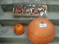 Two rattan pumpkins, large (18" wide) and small (7" wide), on concrete stairs alongside a packaged set of fall-themed miniature village houses and figures