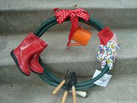 Front view of the 18-inch garden hose wreath decorated with miniature garden tools, red rubber boots, a red and white polka dot bow, floral gloves, and an orange watering can.