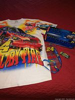 Full set laid out showing T-shirt, both diecast cars, lanyard, and CD case on red surface.