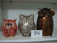 Front view of three owl figurines: small red ceramic tea light holder, medium silver metal owl, large dark metal owl with gold designs.