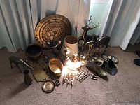 Overview photo of all brass items grouped on carpeted floor including candlesticks, deer figurines, decorative bowls, garrafe figurine, lamp, and other items