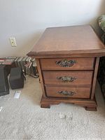 Side view of the solid wood end table showing the top and front with two drawers and metal handles. The carpet and adjacent items give context to size and condition.